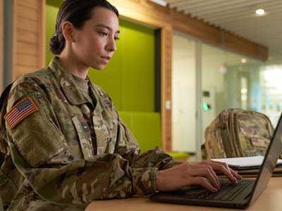 A person in a camouflage military uniform typing on a laptop.