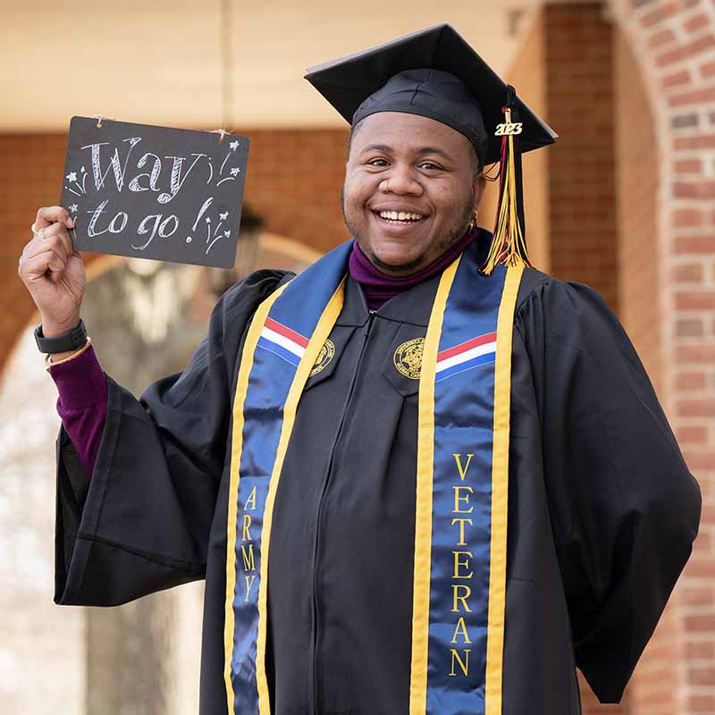 A UMGC graduate in graduation regalia holding a sign that says, "Way to go!"