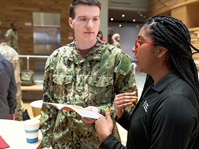 A woman in a UMGC shirt shows a man in military uniform a brochure and speaks with him.
