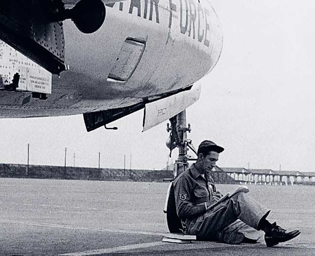 A person in an armed services uniform is resting against the wheel of an Air Force plane.