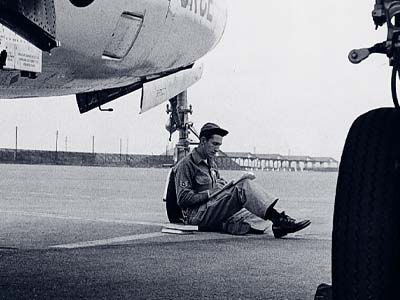 A black and white photo of a person in a military uniform sitting beside a plane.
