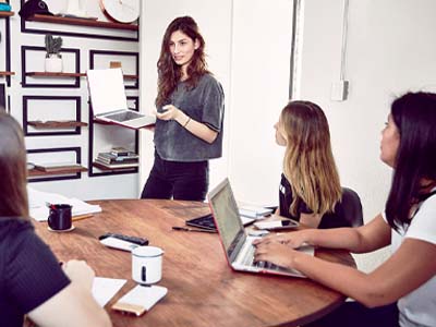 A person holding a laptop standing up while three people sit around a table.