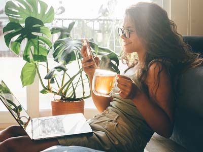 A person smiling while holding a clear mug of tea and holding a cell phone in the other hand with a laptop on their lap.