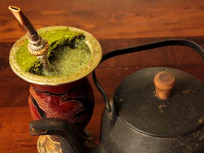 Green yerba mate in a decorative mug next to a tea kettle.