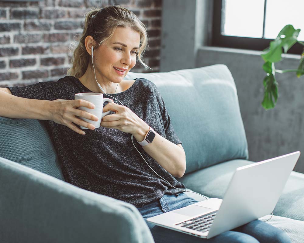 A person holding a mug while wearing ear buds with a laptop on their lap.