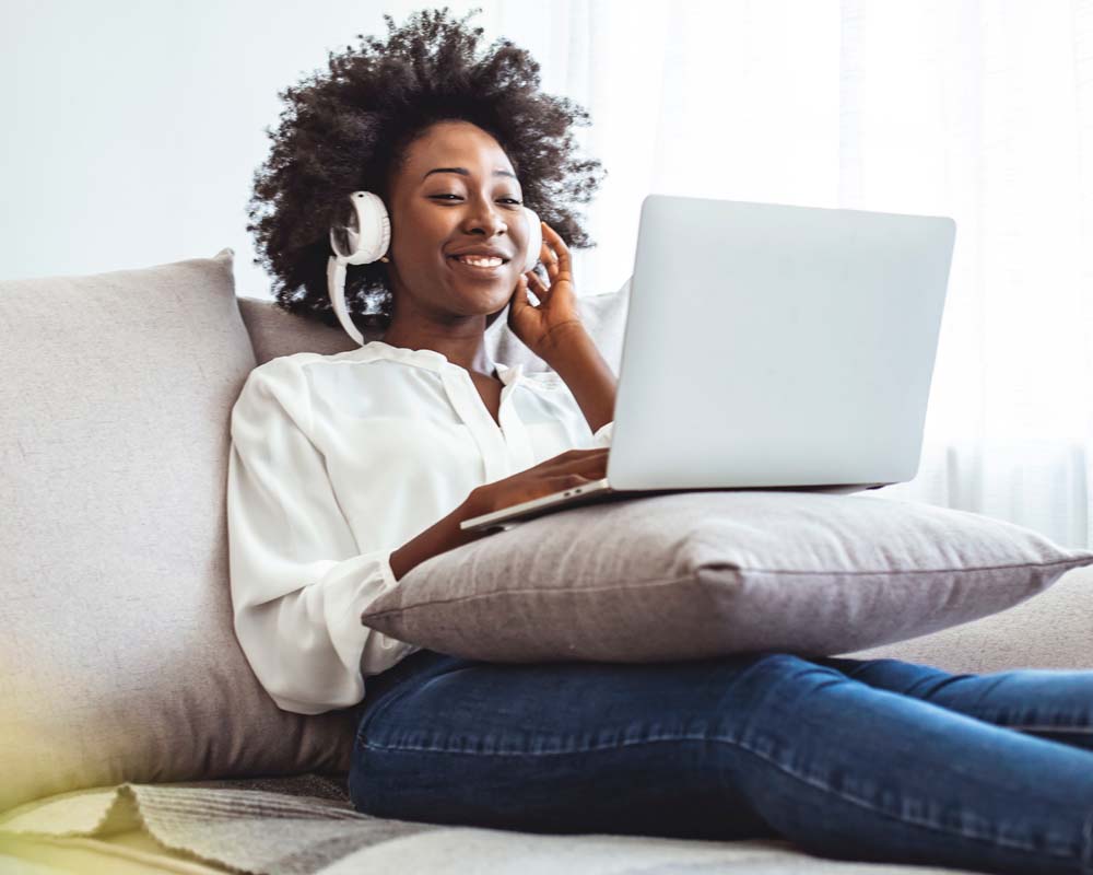 A person wearing headphones sitting on a couch with a laptop, smiling.