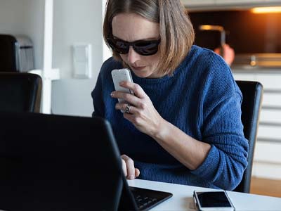 A person with sunglasses near a laptop and a mobile phone, holding a device.