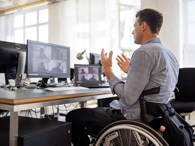 A person in a wheelchair sitting at a desk with multiple monitors, talking on a video conference.