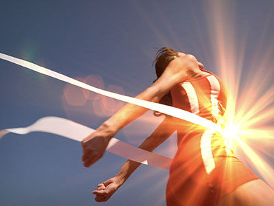 A person breaking through finish line tape with bright sun in the background.