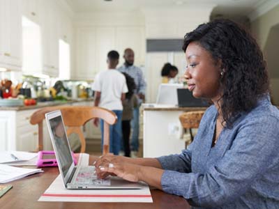 Woman sits at a dinner table and types on her laptop while her family is in the kitchen behind her.