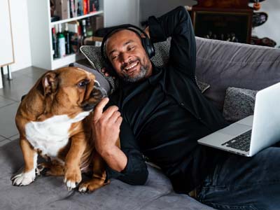 A person with headphones on smiling while lying on a couch with a laptop and a dog.