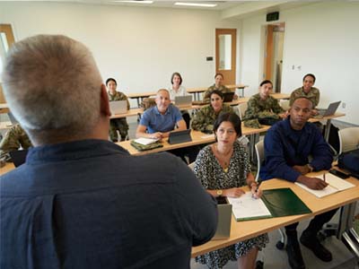 A man stands and talks to a class of adults while the class looks at him and listens.