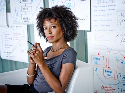 Woman sitting alone in a chair holding a pen in front of an array of hand-written charts hanging on the wall.