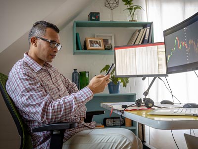 Man sitting alone in a home office viewing his phone in front of a desk with two large computer screens.