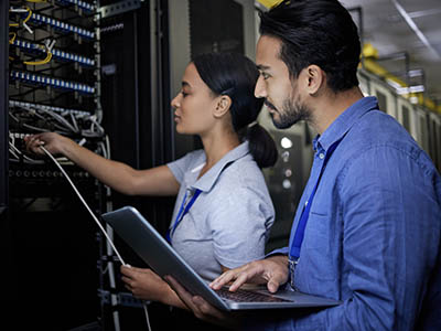 A man holds a laptop and watches a woman plug in different cords in a data center.