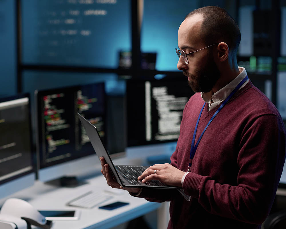 A man wearing a red sweater and key lanyard types on a laptop in an office, with three other computer monitors turned on in the background.
