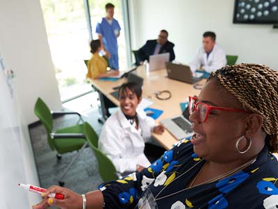 A group of people sit in a conference room. Four people chat while writing notes and one writes on a whiteboard while another watches.