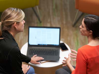 Two people sitting and talking while one person types on a laptop.