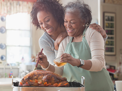 Elderly mother smiling and basting a turkey while her adult daughter leans in close and smiles.