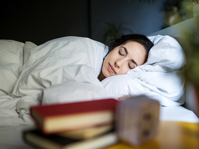 Woman in bed sleeping on her side with a books on a nightstand in the foreground.