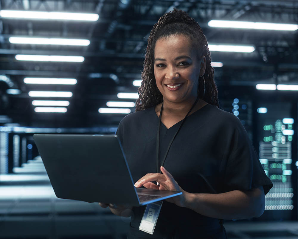 A woman holding a laptop and smiling at the camera while working inside a data center.