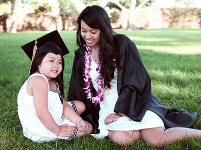 A child wearing a graduation cap sitting on the grass next to a person in a graduation gown.