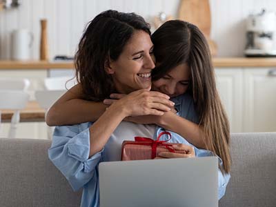 A child giving a wrapped gift and hugging a person on a laptop.