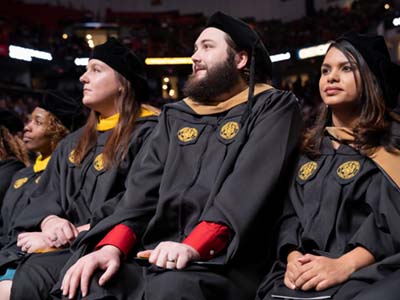 Three graduate students in caps and gowns at their commencement ceremony sit and listen to a speaker on stage