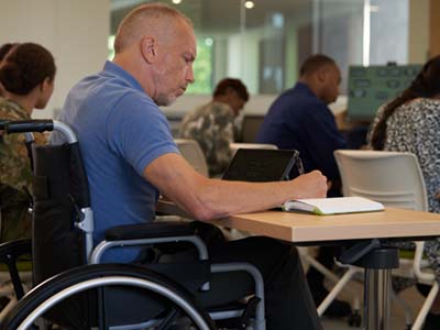 Man in a wheelchair sits at a desk and reads a textbook in a class with others.