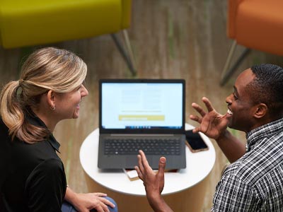 Two people sit and talk to each other in front of a shared laptop.