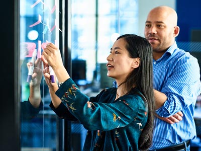 A woman puts a note on a wall while a man stands next to her watching.