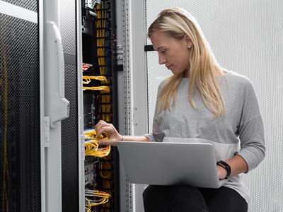 A woman sits with a laptop in front of a large mainframe computer and plugs in a wire.