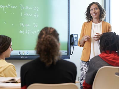 A woman standing next to a large wall-mounted screen in front of three seated students