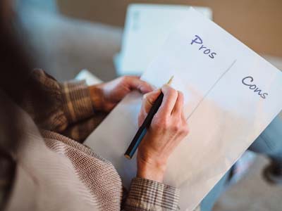 A person's left hand holding a paper with columns for Pros and Cons in their lap and their right hand holding a writing implement to the paper