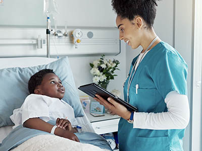 A person in scrubs holding a digital tablet and talking to a child in a hospital bed.