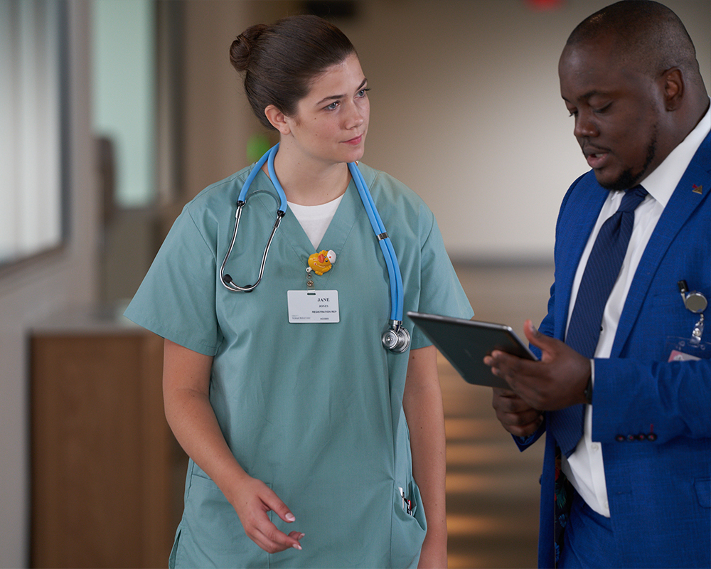 A person in medical scrubs talking to another person with a digital tablet.