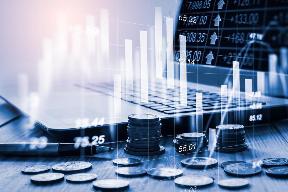 A stock image of a computer with coins beside it. A graph is overlaid on top of the coins and laptop.