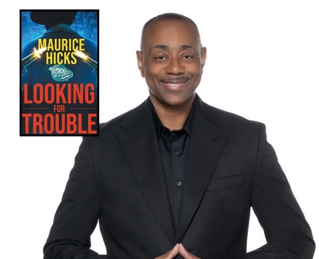 Maurice Hicks looks at the camera while wearing a suit that is black with a black shirt. He is smiling. In the top corner, the cover of his book, Looking for Trouble, is also shown, featuring a detective looking away and police badge plus guns.