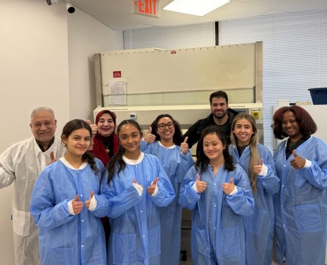 Six UMGC biotechnology master’s students in blue lab gowns pose with IPS leadership at the company’s facility in Rockville, Maryland