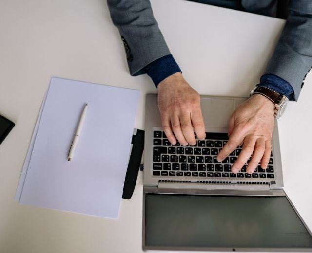Business person’s hands typing on a laptop, with pen and paper nearby. 