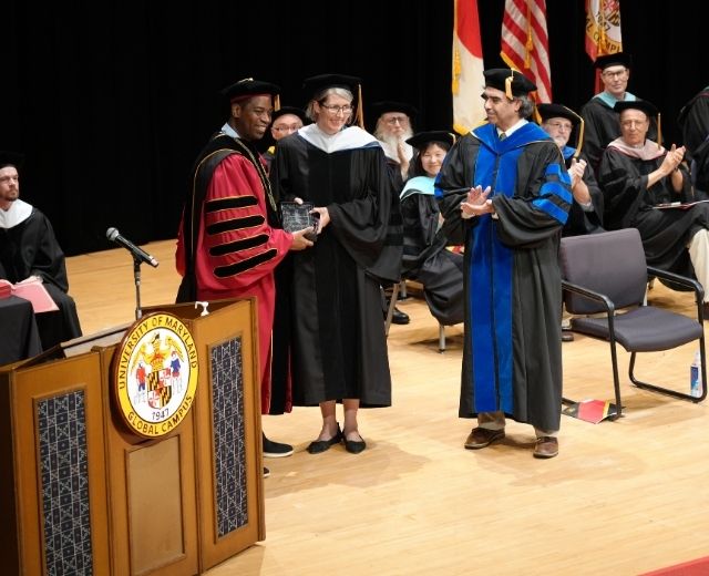 President Fowler presents a Drazek Award to Michelle Fukuyama, PhD, at the UMGC in Asia commencement ceremony in Okinawa, Japan on April 18, 2026, on stage with several other UMGC faculty members.