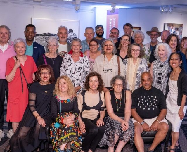 A group of about 24 artists smile in a group photo with UMGC President Fowler in the gallery space at the 6th Biennial Maryland Regional Juried Art Exhibition opening reception on June 7, 2026. 