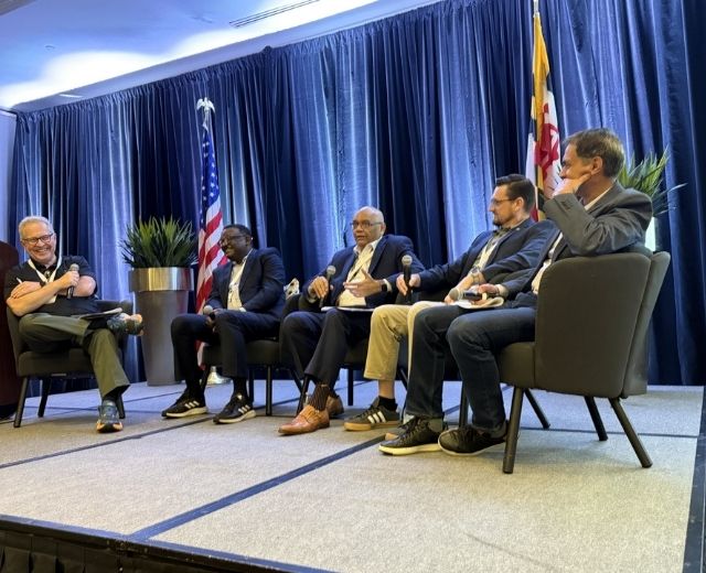 Five men are shown speaking on a stage for a panel discussion on AI in higher education. 