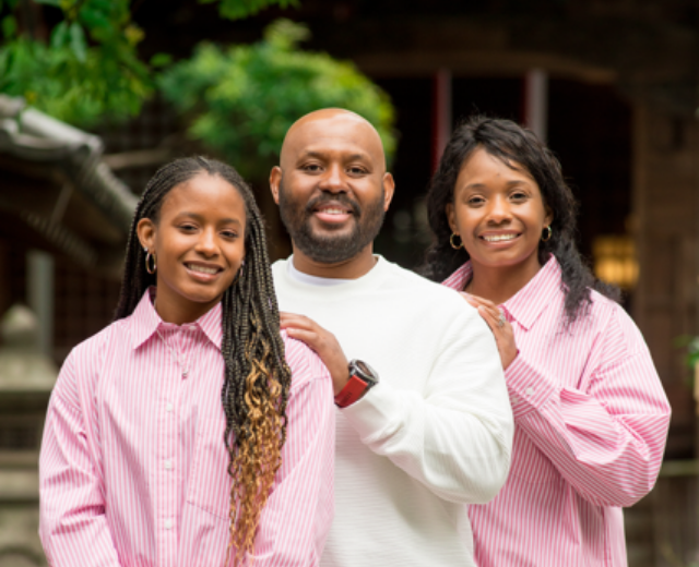  Three members of the Calliste family. Daughter, husband, and wife smile in a family photo. The women are wearing matching pink shirts.