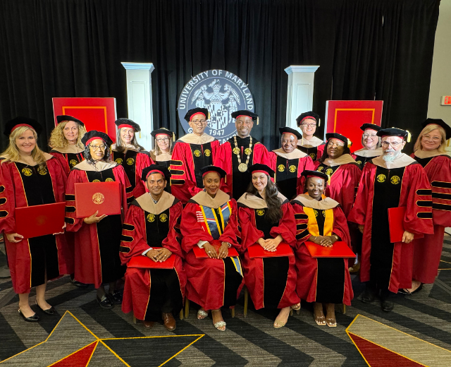 Sixteen doctoral graduates in red robes and black hats pose for a group photo with UMGC President Greg Fowler, PhD, also in red regalia. A UMGC seal is in the background.
