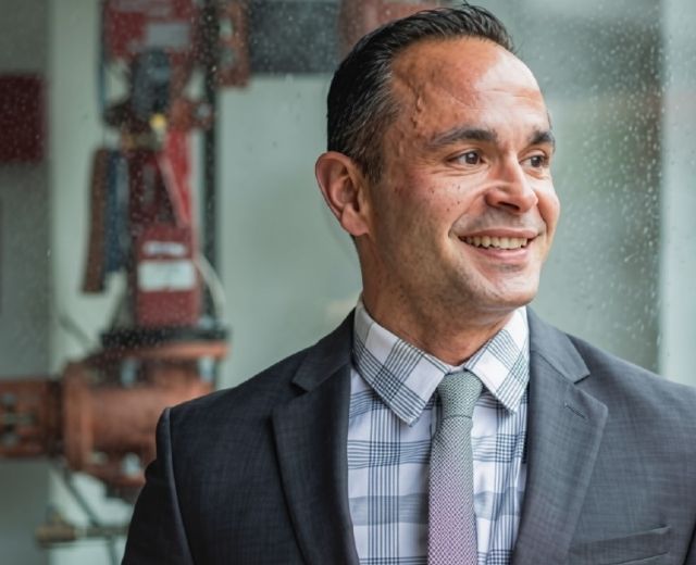 Dan Fernandez in a suit and tie, with red pipes in the background.