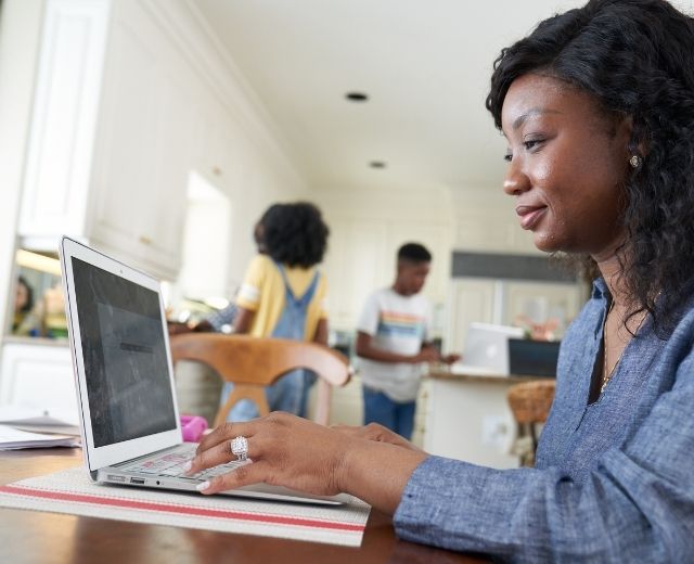 A woman works on a laptop near her children doing homework in the kitchen. 