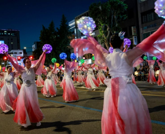 Women in pink and white dresses dance in the street during a parade. They are holding lit ball lanterns in their hands above their heads.