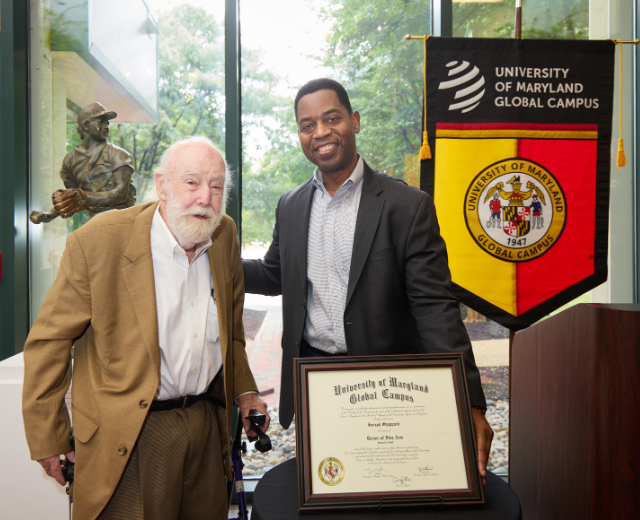 Joseph Sheppard stands with Dr. Fowler behind a framed degree at the Leroy Merritt Center for the Art of Joseph Sheppard in Adelphi, Maryland. A banner with the University of Maryland Global Campus seal is in the background.
