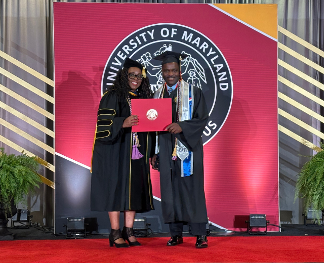 Hannah Anderson Hughes on stage in regalia presents a red folder to a graduate also wearing a cap and gown.